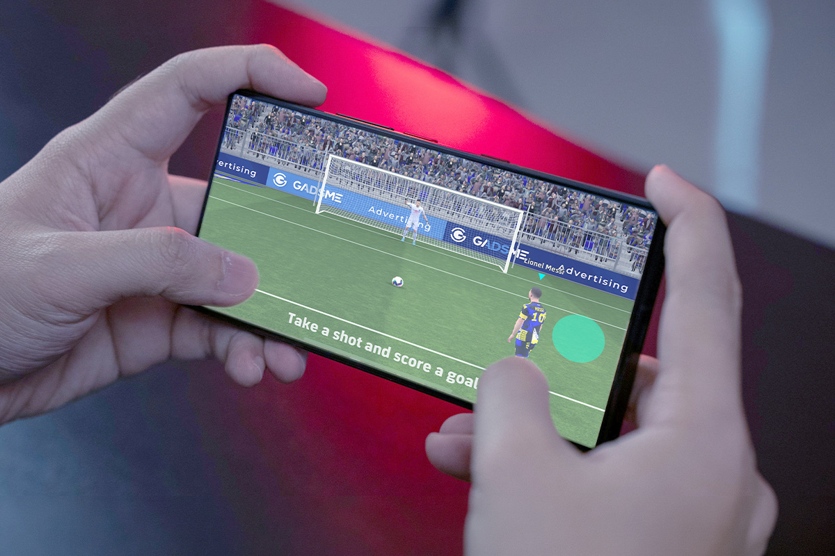 Hands holding a phone running a football game with Gadsme boards behind the goal and a prompt to take a shot